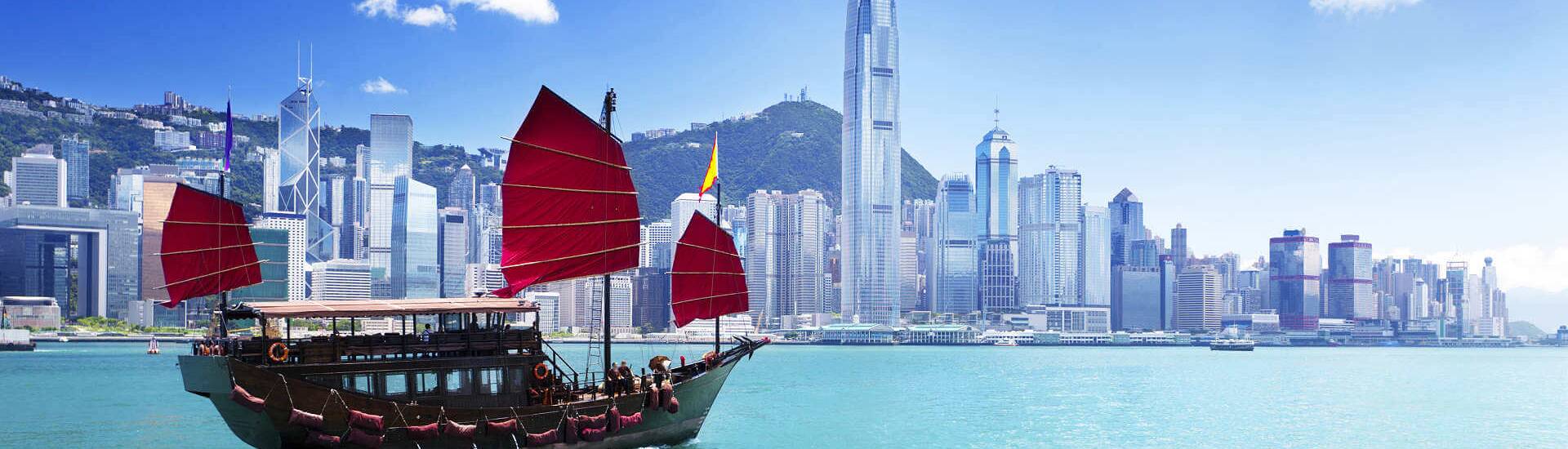 Hongkong mit Wolkenkratzern mit Berglandschaft und im Vordergrund eine Dschunke.