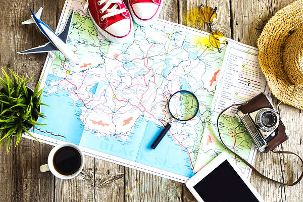 Weltkarte mit verschiedenen Reiseutensilien wie einer Kamera, einem Vergrößerungsglas, einem Tablet, einem Hut und einem Paar Schuhen.