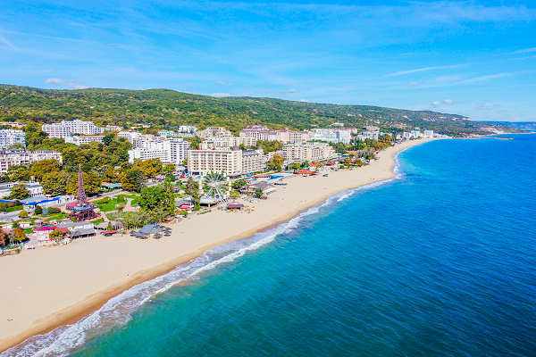 Blick auf den Goldstrand in Bulgarien mit goldenem Sand, klarem Wasser und blauen Sonnenschirmen unter einem strahlend blauen Himmel.