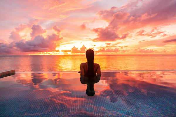 Frau im Pool beobachtet einen atemberaubenden Sonnenuntergang auf Santorin, Griechenland, mit dem tiefblauen Meer im Hintergrund.